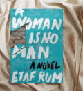 The book "A Woman is No Man" is laying on crumpled beige fabric.